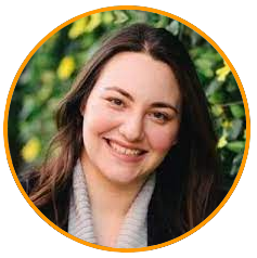 A person with long brown hair wearing a light gray scarf, standing in front of green foliage.