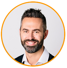 Professional headshot of a man with a beard and short dark hair, wearing a white shirt and dark jacket, smiling in front of a light background inside an orange circular border.