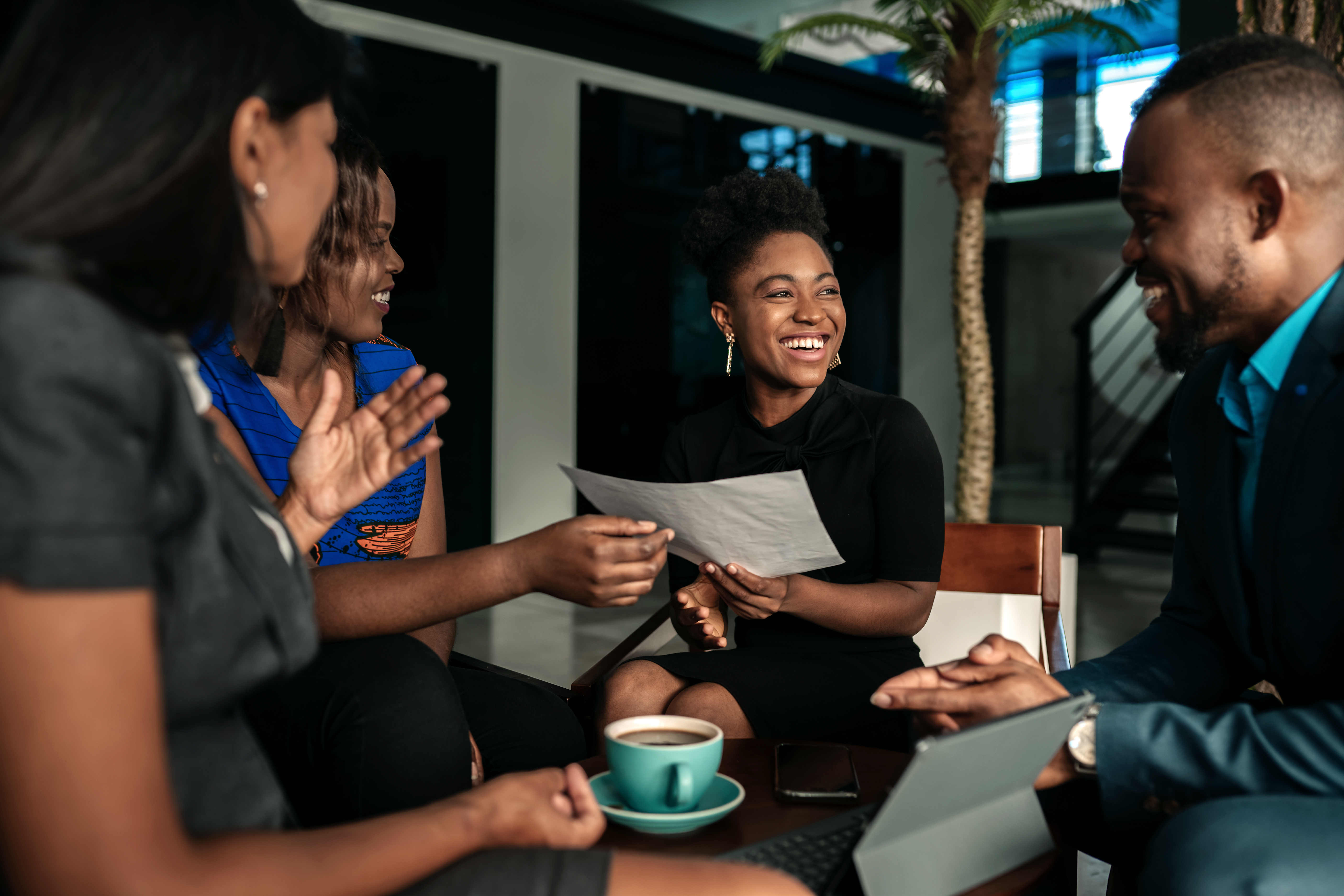 A group of professionally dressed colleagues having a casual and happy meeting around a small table with coffee, documents, and a laptop in a modern office setting.