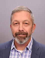 Headshot of a business professional with a gray beard, wearing a blue blazer and checked shirt, against a neutral background.
