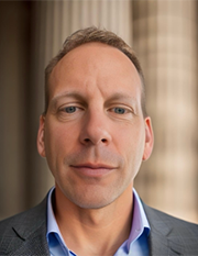 A professional portrait of a middle-aged man in a suit standing in front of columns, suitable for use in a business or corporate setting.