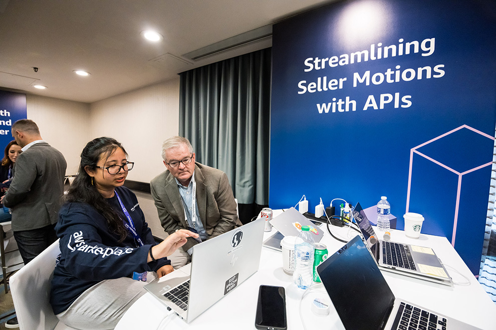Two participants engage in a discussion and demonstration at a table with laptops during the AWS Seller Conference, in front of a sign that reads 'Streamlining Seller Motions with APIs.'