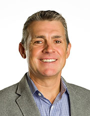 Professional headshot of a smiling man wearing a gray blazer and blue shirt, posed in front of a white background.