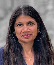 Portrait photo of a woman with long dark hair, wearing hoop earrings and a navy blouse, posing in front of a blurred gray background.