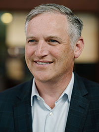 A portrait photo of a smiling man with short gray hair, wearing a blue blazer and collared shirt, taken at a conference setting.