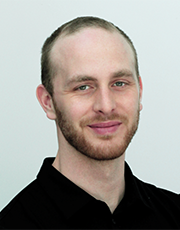 Professional headshot of a man with light skin, short hair, and a trimmed beard, wearing a black shirt, posed against a plain light background.