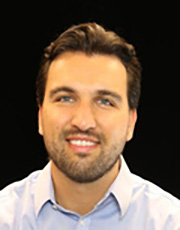 Professional headshot of a man with short dark hair and a beard, wearing a light blue collared shirt, posed against a dark background.
