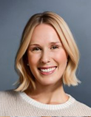 Portrait of a smiling woman with short blonde hair, wearing a light-colored sweater against a gray background.