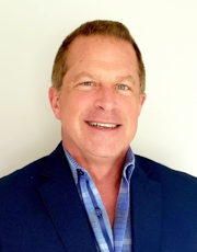 A professional headshot of a man wearing a blue blazer and light blue shirt, smiling against a plain background.