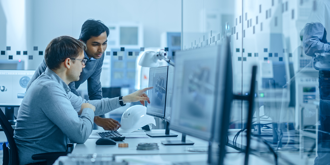 Two people collaborating at a desk with computer monitors displaying 3D models in a modern office or lab setting.