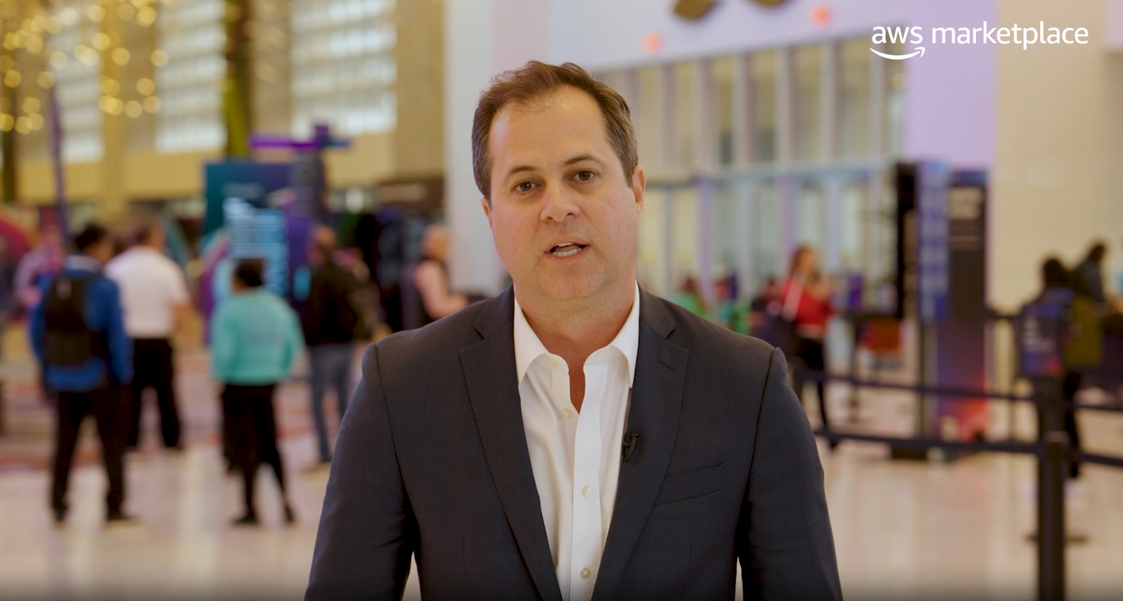 A professionally dressed individual stands in focus in front of a blurred background at an event, with the AWS Marketplace logo visible in the upper right corner.