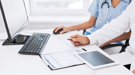 Two healthcare professionals reviewing documents and a tablet at a desk with a computer.