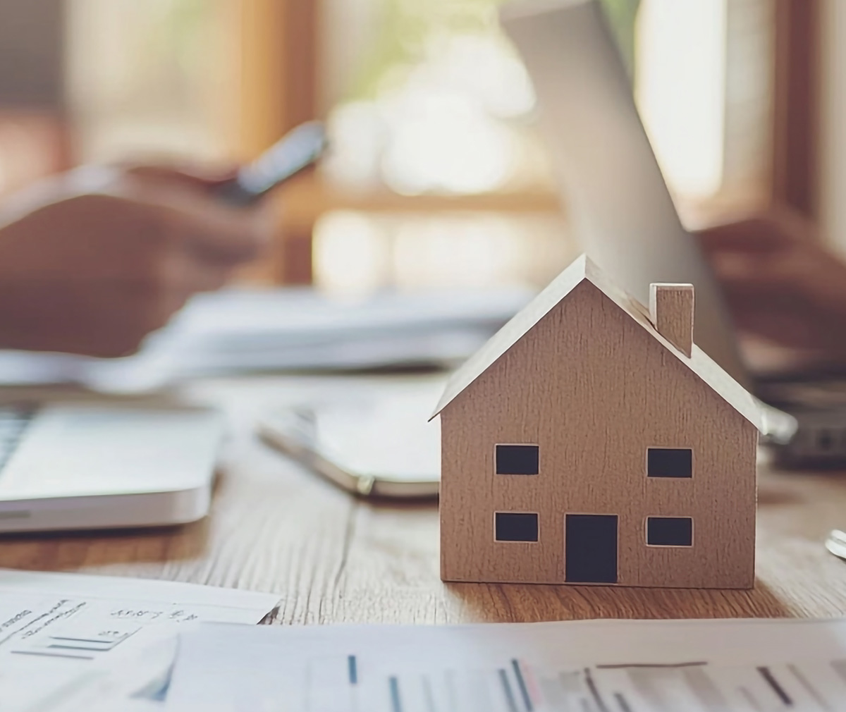 A small wooden house model on a desk surrounded by papers, with blurred hands using a phone and laptop in the background.