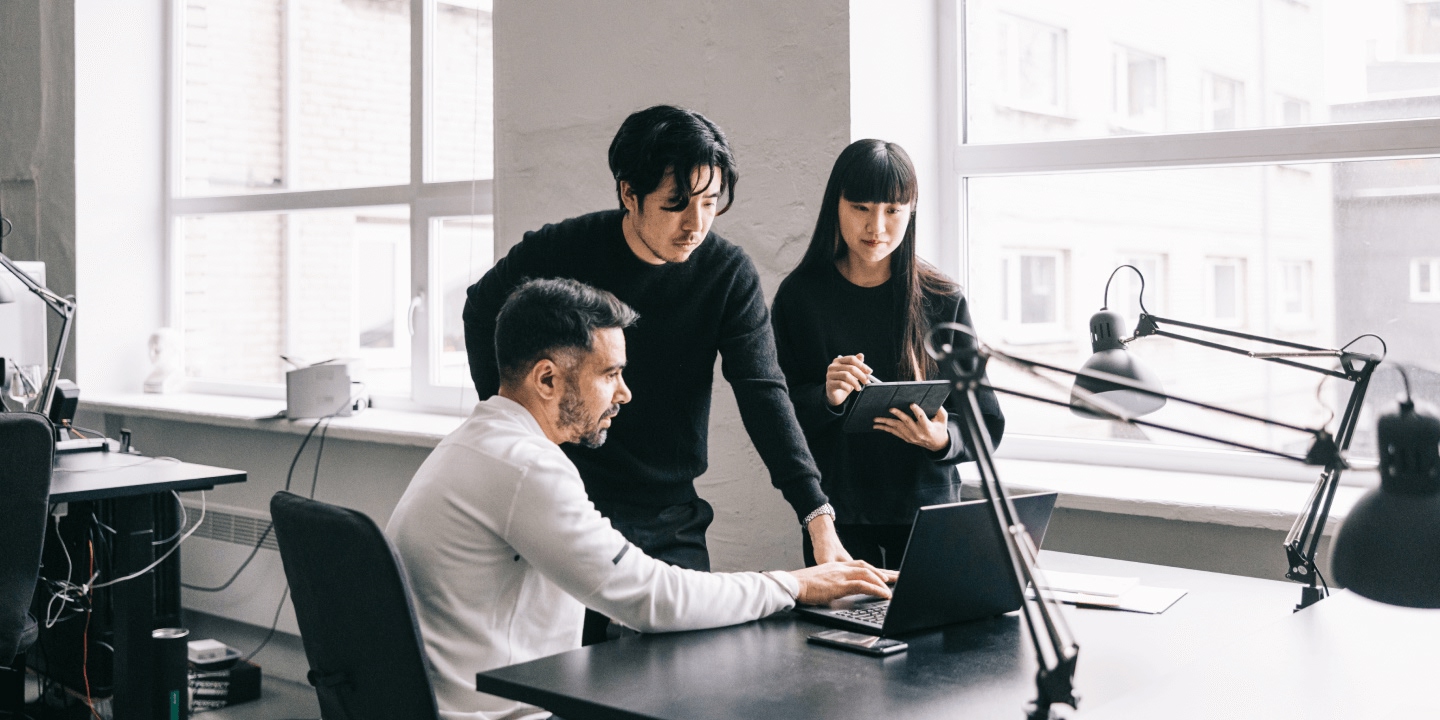 Three content creators collaborate during podcast recording in bright modern office. One seated at laptop with professional microphone, two standing with tablet. Large windows overlook urban cityscape with natural lighting.