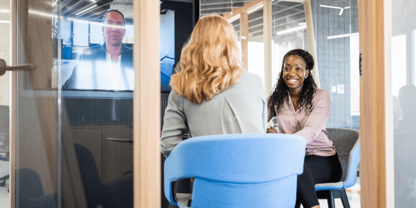 Three people meet in glass-walled conference room with man visible on video display screen, demonstrating virtual collaboration in modern office.