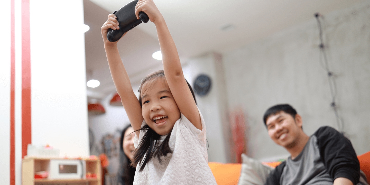 Young child joyfully holding black gaming controller overhead in modern indoor recreation facility, with other children and adult supervisor visible in background, wearing light gray t-shirt.