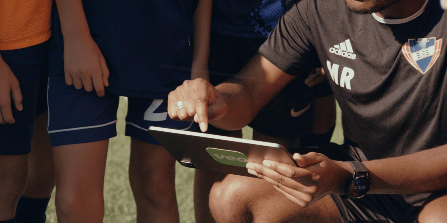 Sports coach wearing Adidas shirt demonstrates tactical information on tablet device to team members gathered around during outdoor training session, showing digital strategy planning in action.