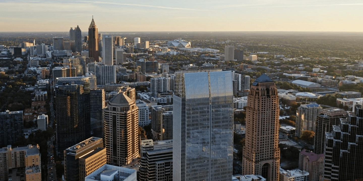 Atlanta downtown skyline aerial photograph during golden hour. Tall Art Deco spired building anchors cluster of modern glass skyscrapers and office towers. Extensive tree coverage surrounds urban core extending to horizon. Clear sky with warm afternoon lighting.