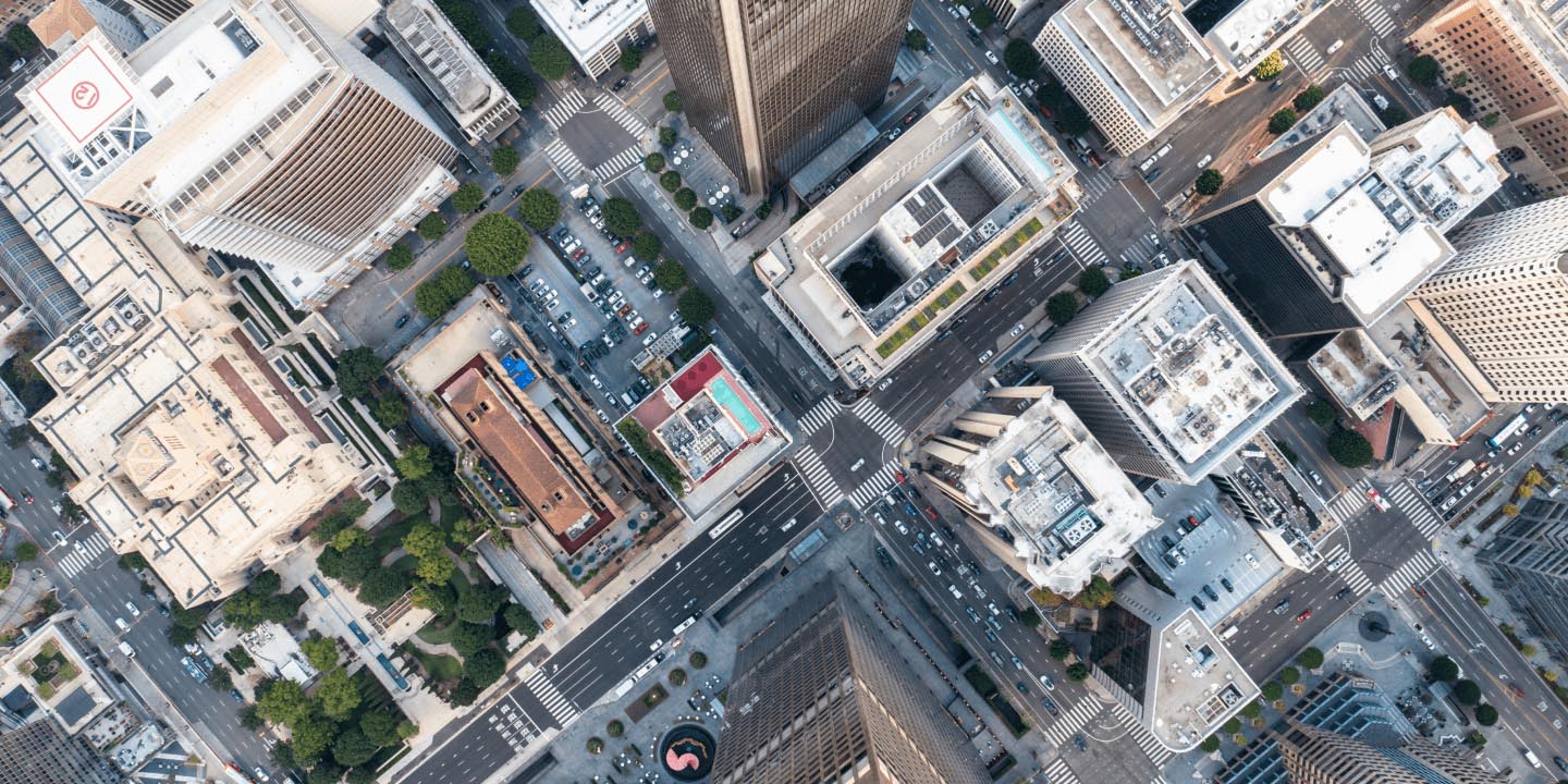 Aerial view of dense urban city block with mixed-use buildings, high-rise structures of varying heights, pedestrian crosswalks, parked vehicles, street grid pattern, landscaping and trees throughout.