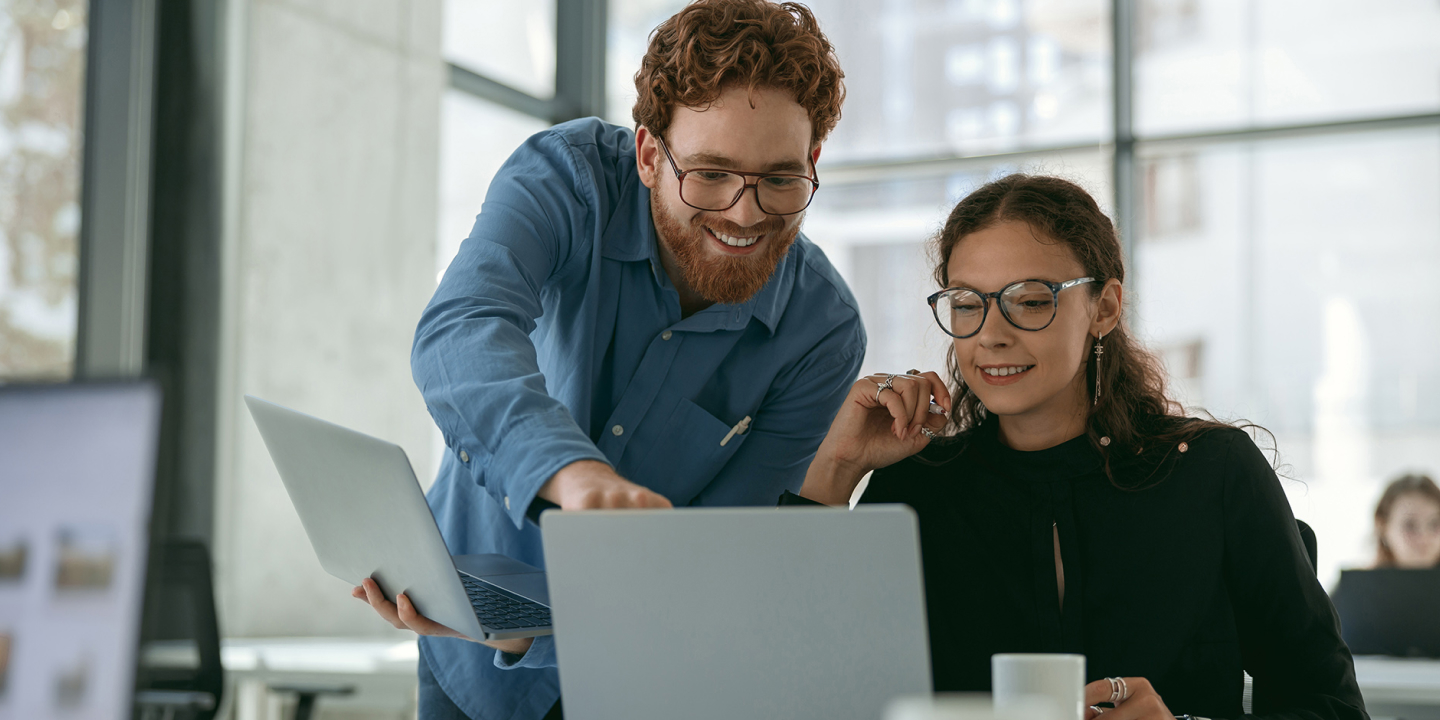 The main subjects are a man and a woman who are working on a laptop. They are in an office environment, with large windows in the background. The man is wearing a blue shirt and has a beard, while the woman is wearing a black blouse and has curly hair. They are both wearing glasses. The laptop is silver, and the background features a modern office with glass windows.