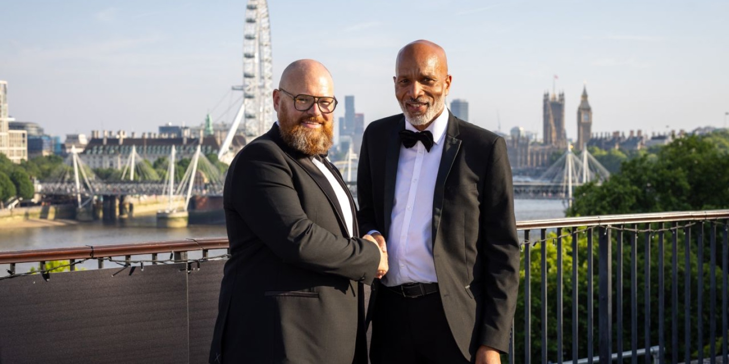 Two men in black tuxedos and bow ties shake hands outside with a river view.