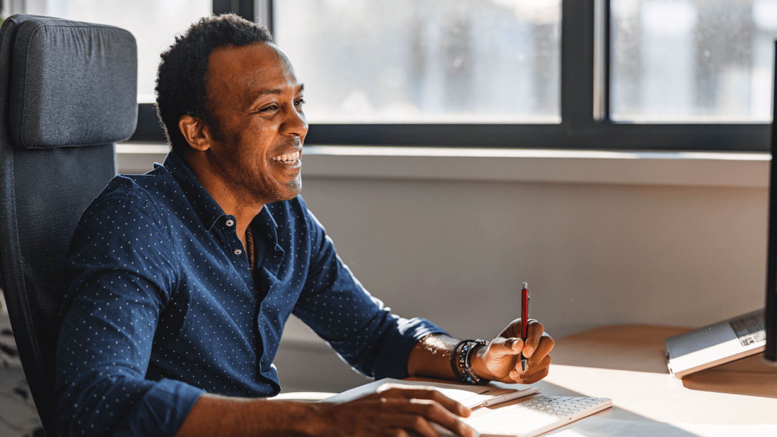 Man smiling at a desk with a laptop and notebook beside a sunlit window