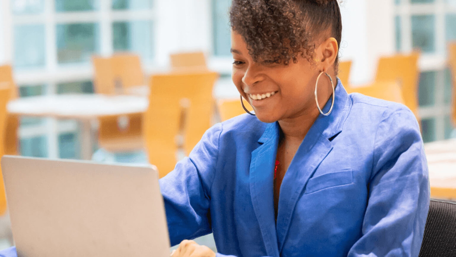 A woman with curly hair is seated on a chair, smiling, and using a laptop. She is wearing a blue shirt and silver hoop earrings. The background shows several empty tables and chairs, suggesting she is in a cafeteria or a similar setting. The setting is well-lit, with natural light coming through the windows.