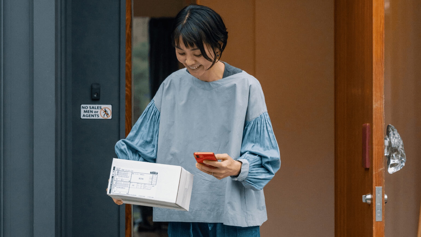 An Asian woman, dressed in blue attire, is standing at the entrance of a room, holding a white box in one hand and a red cell phone in the other. She is smiling and appears to be posing for a photograph.