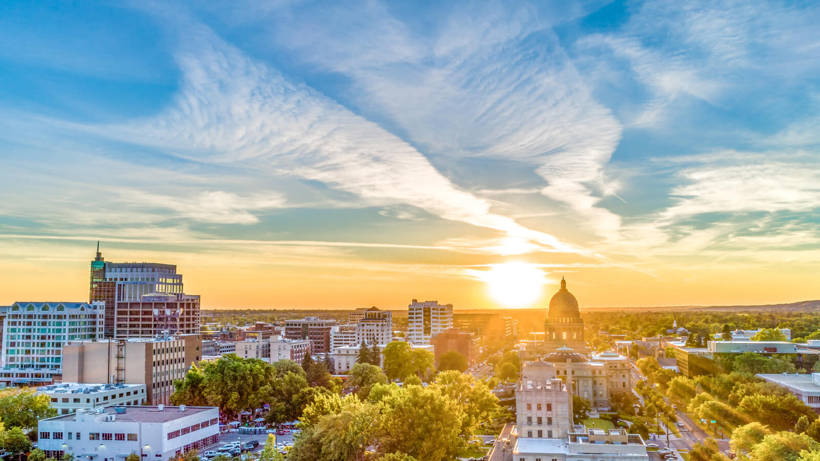 A city skyline at sunset, with the sun setting behind the clouds in the sky. The sun is casting a warm glow on the buildings and trees below. The city is surrounded by trees and has a mix of modern and traditional architecture.