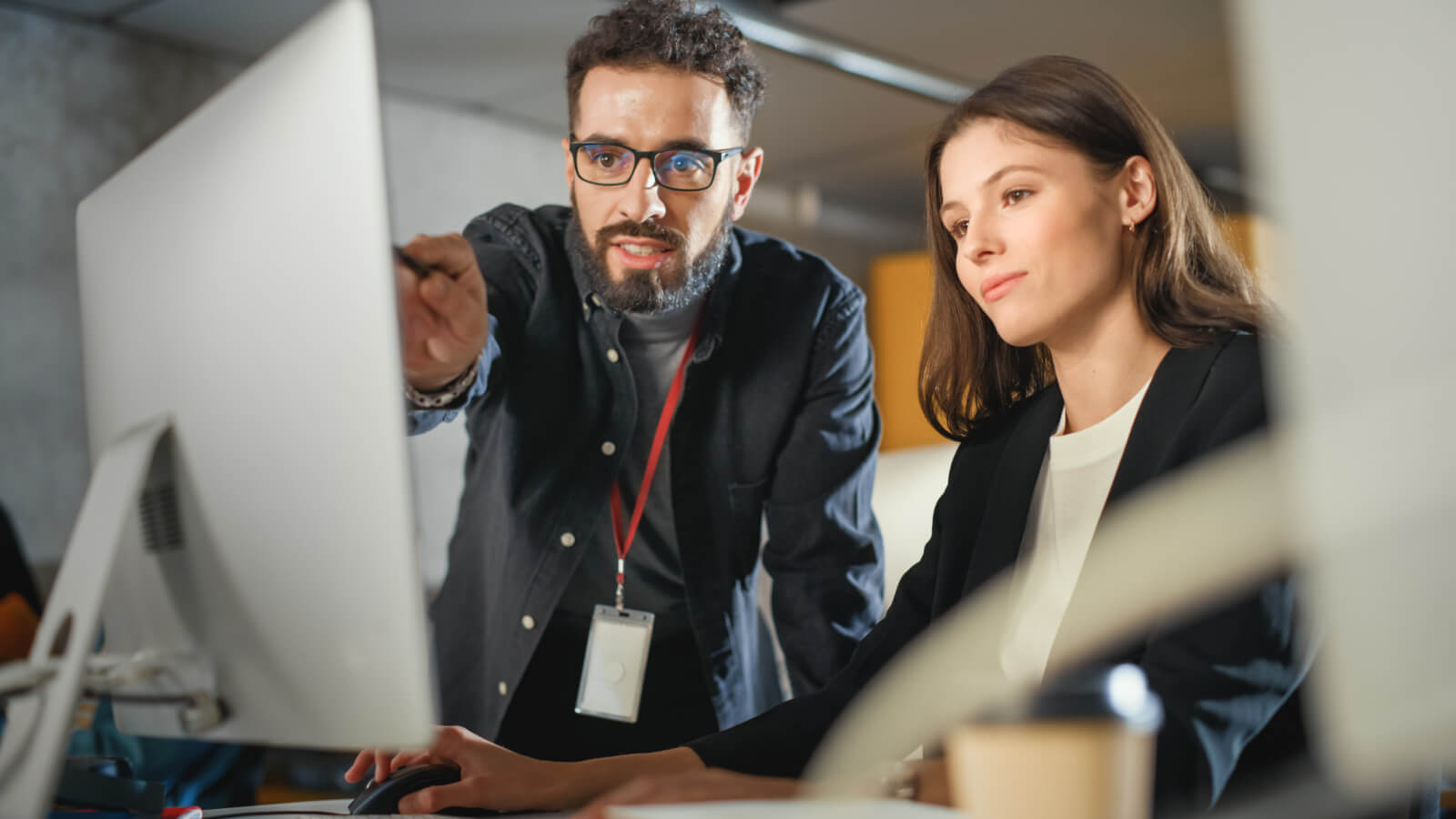 The main subjects are a man and a woman in an office. The man is pointing at the monitor, while the woman is typing on the keyboard. The setting is an office with computers, desks, and office equipment. The man is wearing a black shirt, while the woman is wearing a black blazer over a white blouse. The composition is centered on the interaction between the man and woman.