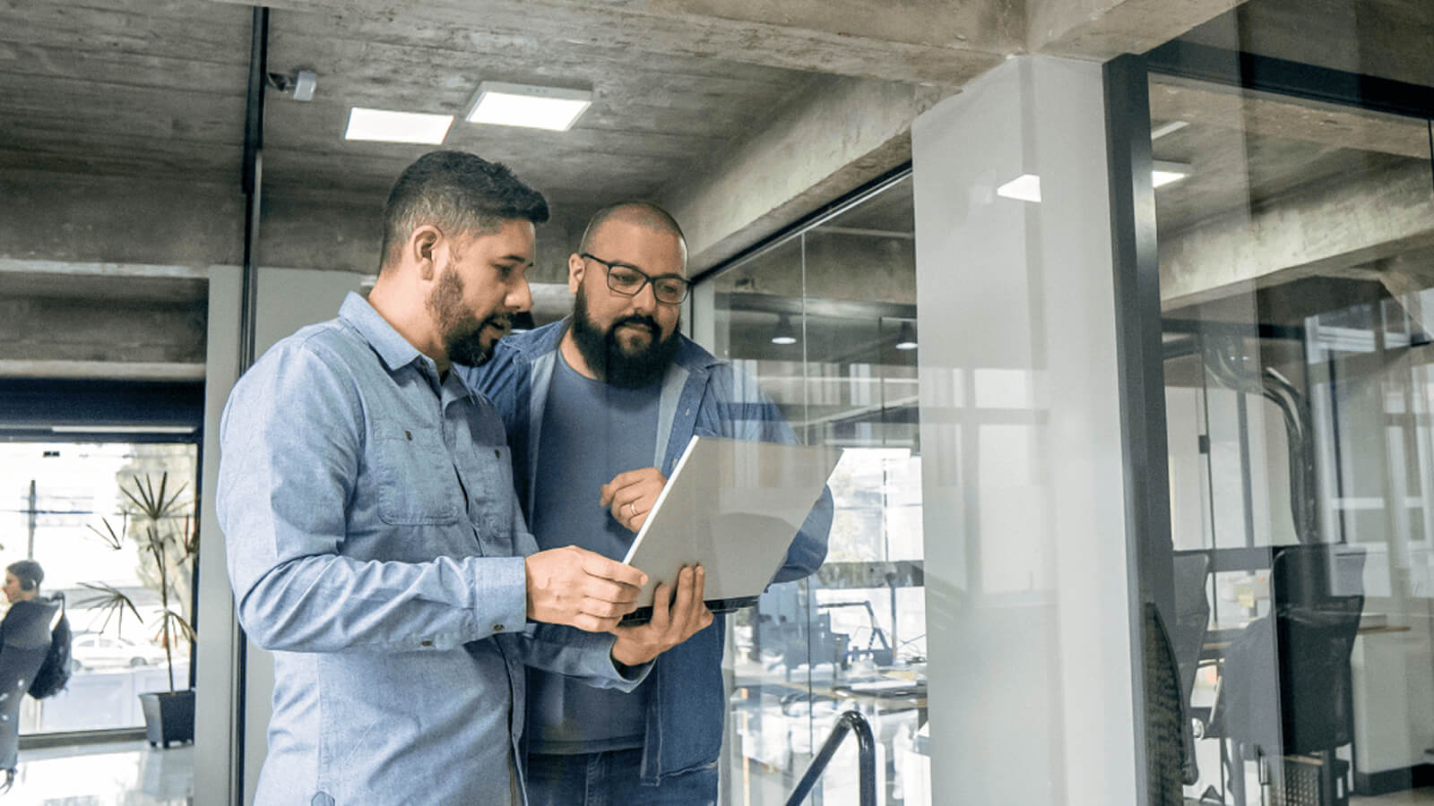 Two men are standing in a modern office setting, one holding a tablet, both looking at it. The office has a sleek glass wall, contemporary furniture, and a minimalistic design. The dominant colors are neutral, with a hint of blue in the men's shirts. The composition focuses on the interaction between the two men, highlighting their collaboration and the use of technology in the workplace.