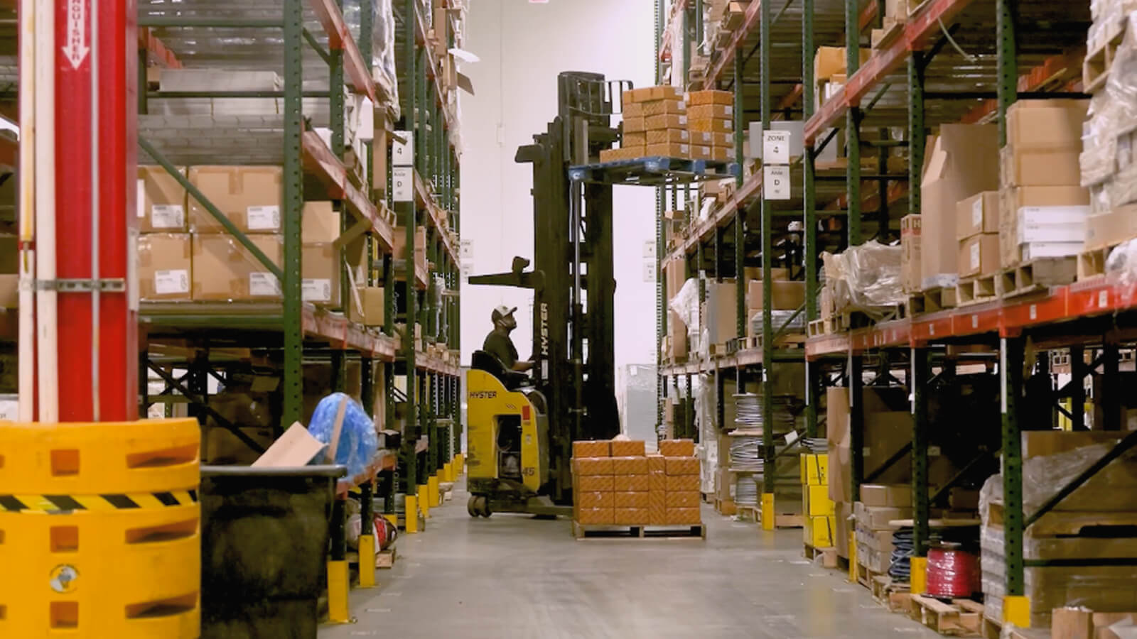 Inside a warehouse, a man is operating a forklift, which is lifting a pallet of boxes. The warehouse is filled with shelves stocked with various items, and the floor is lined with yellow safety barriers. The setting appears well-organized, with clear pathways and efficient use of space.
