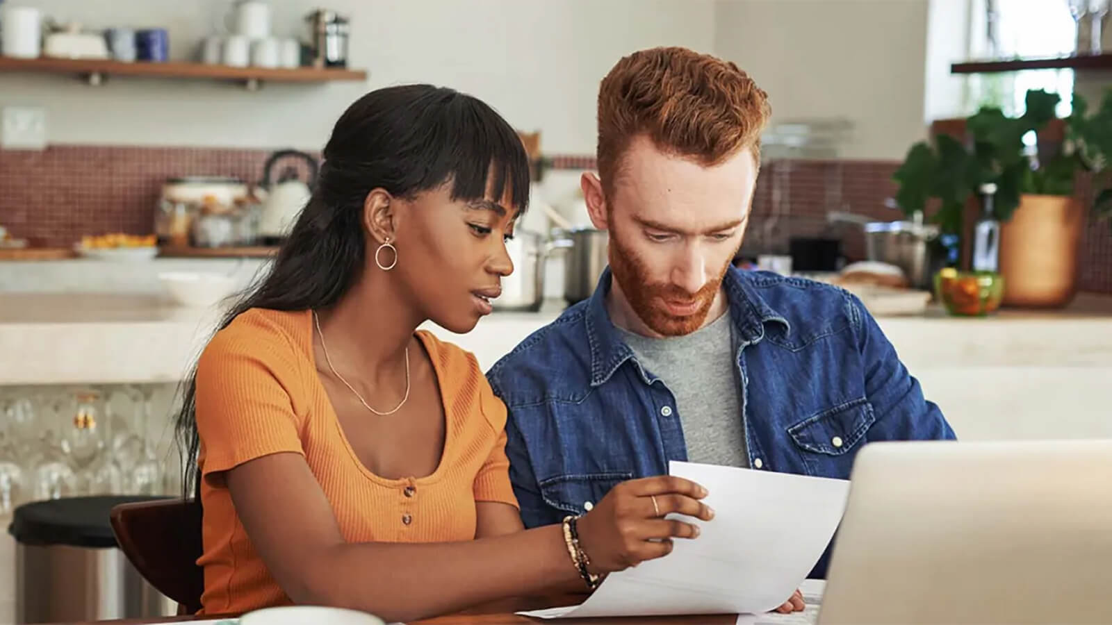 A man and a woman are sitting at a kitchen table, looking at documents. The man is wearing a blue denim jacket, while the woman is wearing an orange top. The kitchen has a countertop with various items, a shelf, and a plant. There is also a laptop on the table. The lighting is warm and soft.