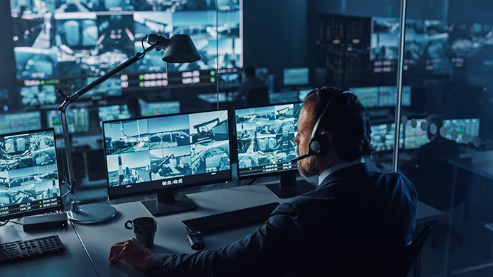 Male Officer Works on a Computer with Surveillance CCTV Video in a Harbour Monitoring Center with Multiple Cameras on a Big Digital Screen. Employees Sit in Front of Displays with Big Data.