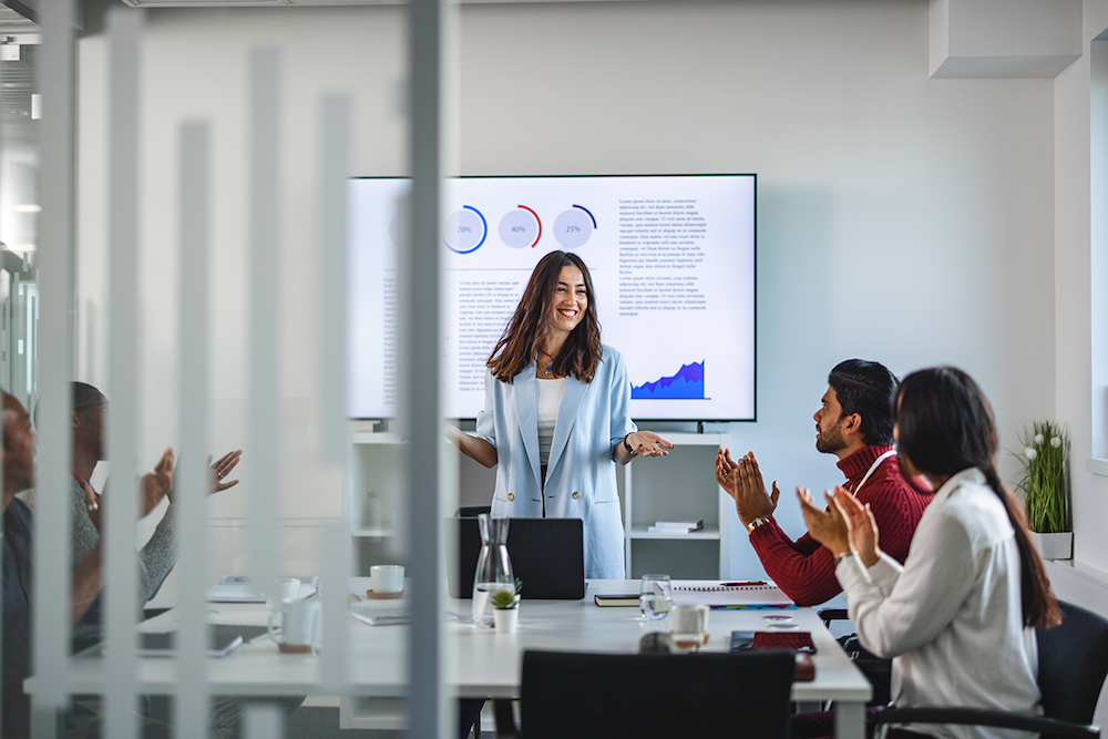 woman standing up in a conference room and two people sitting at a table clapping