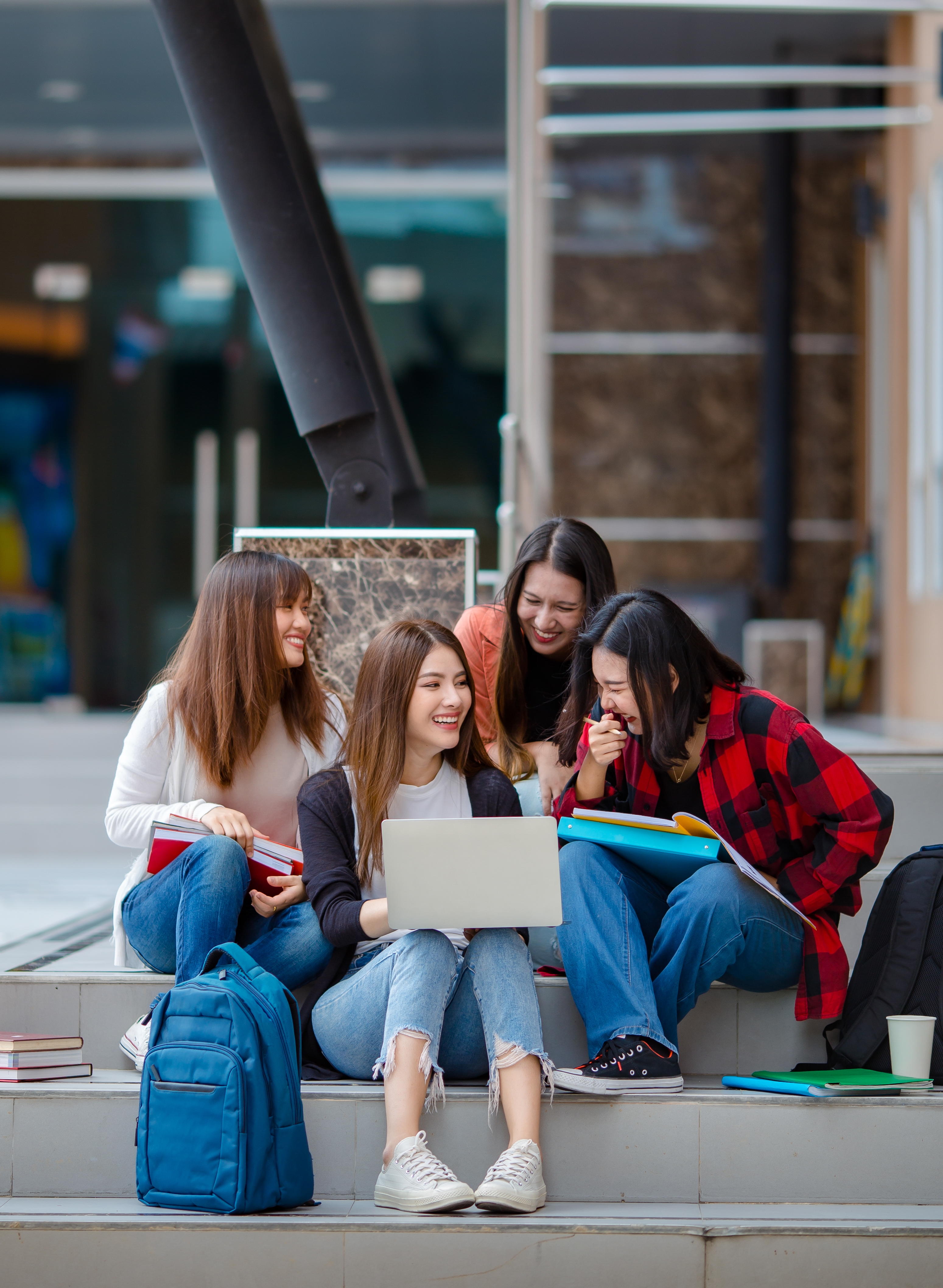 A group of four women sit outside looking at a laptop screen and laughing.