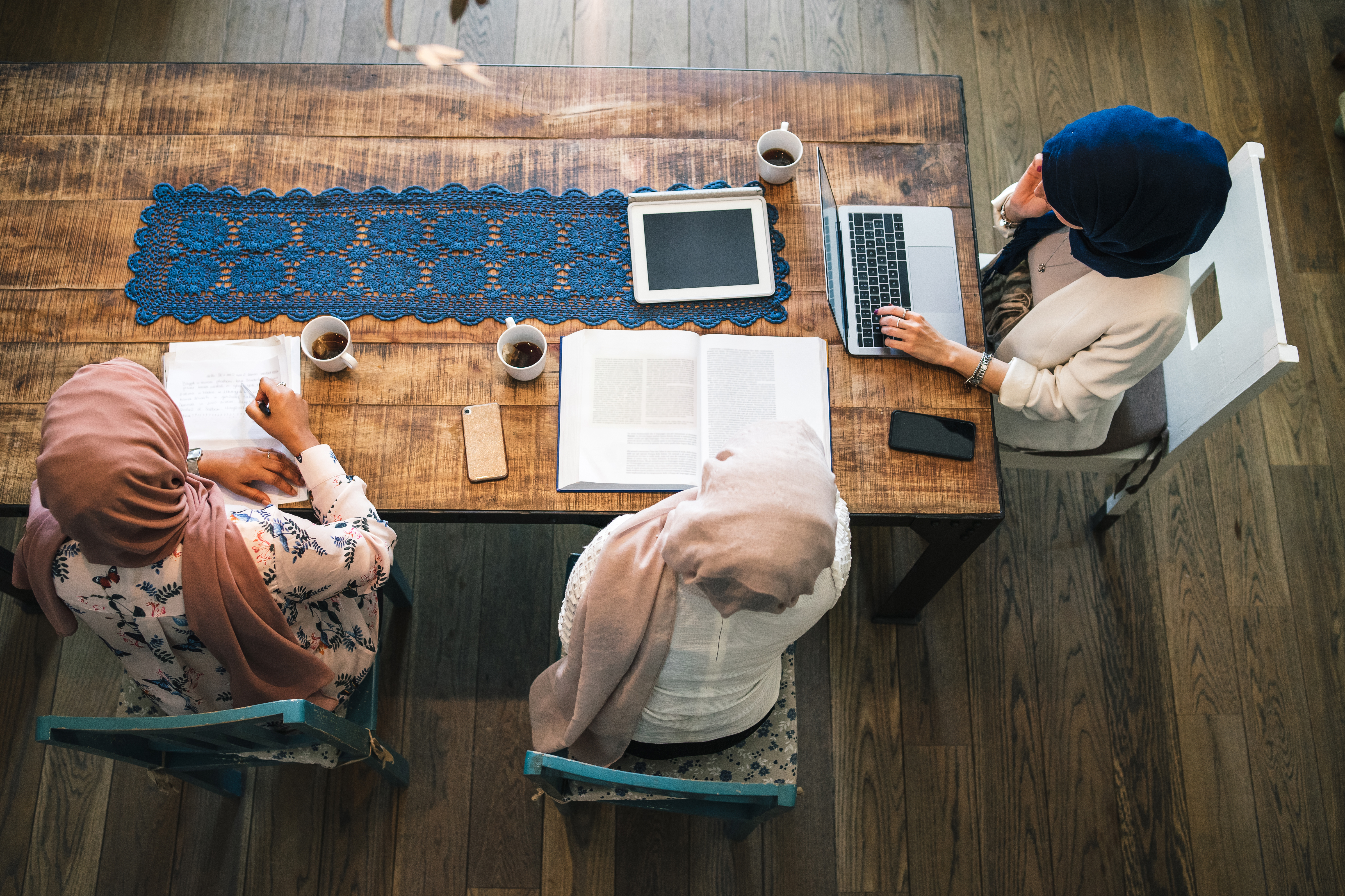 A group of three women sit at a table reading books and open laptops as if studying.