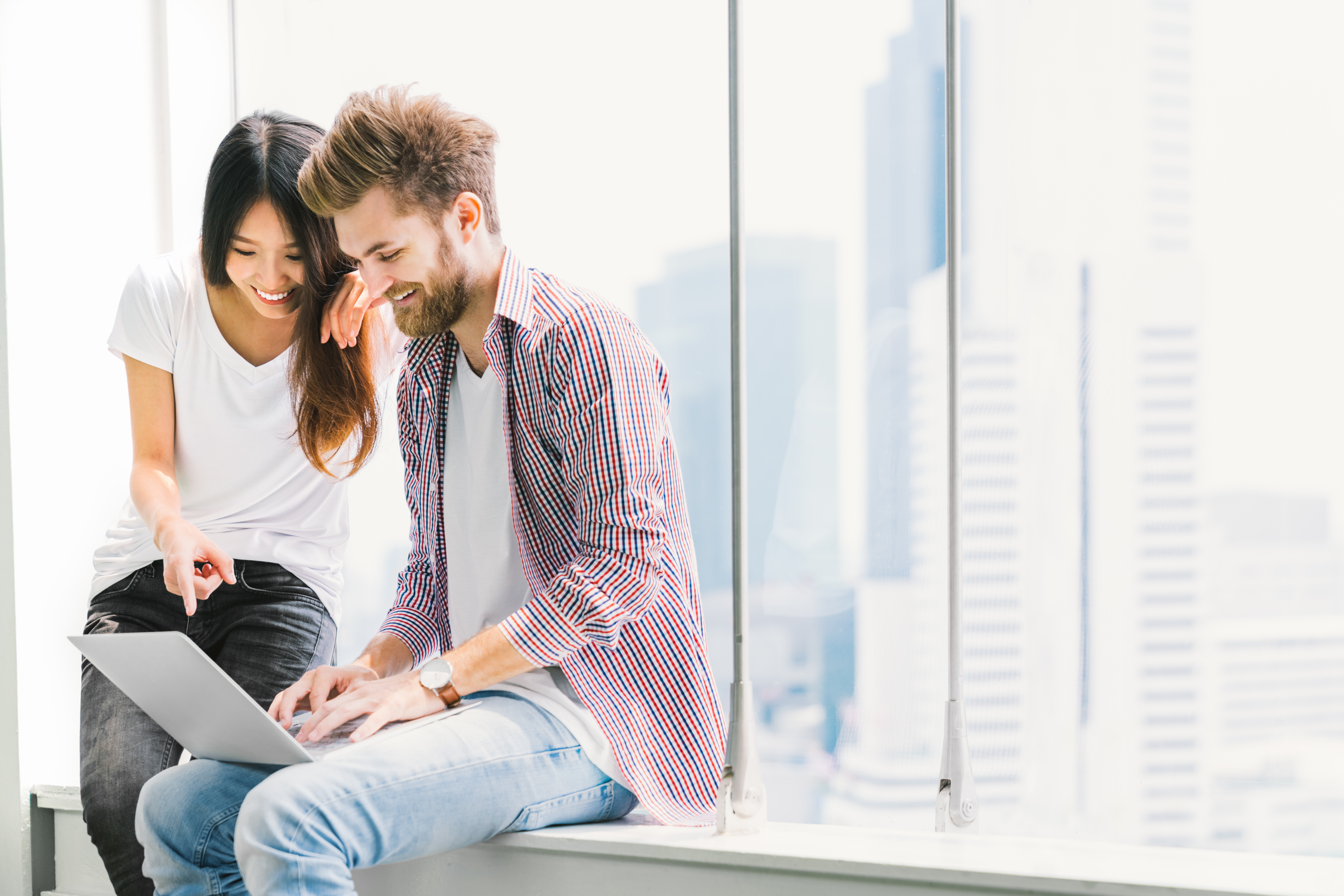 A man and a woman sit together in front of window of an office building, pointing at a laptop screen and look to be in discussion.