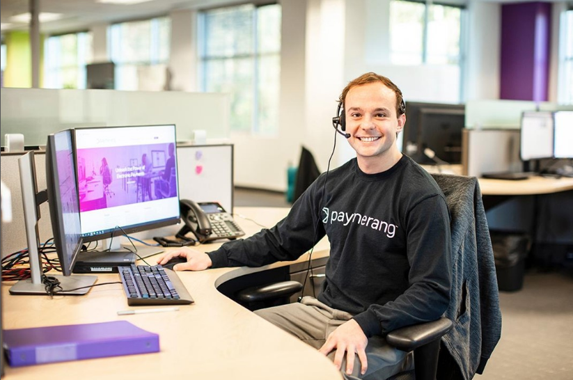 Paymerang employee sitting at a desk smiling at camera with computers in the background.