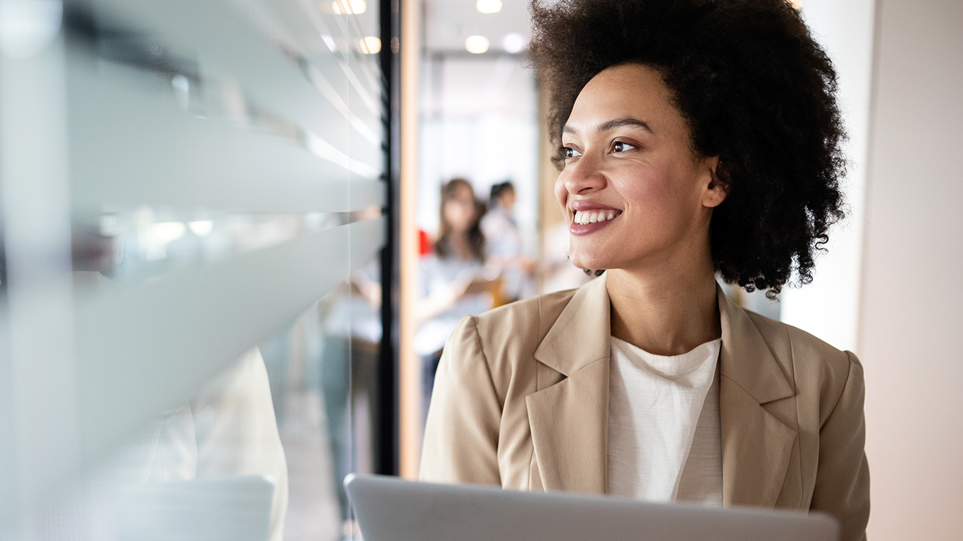 A woman smiling in a hallway and holding a laptop while learning how to deliver professional quality media services using AWS Media Services.